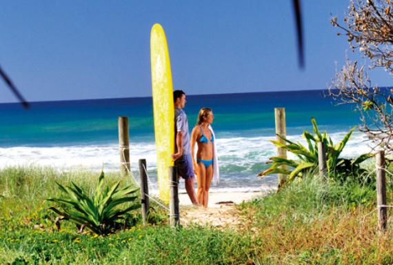 man holding up yellow long surf board with woman in blue bikini standing on the beach