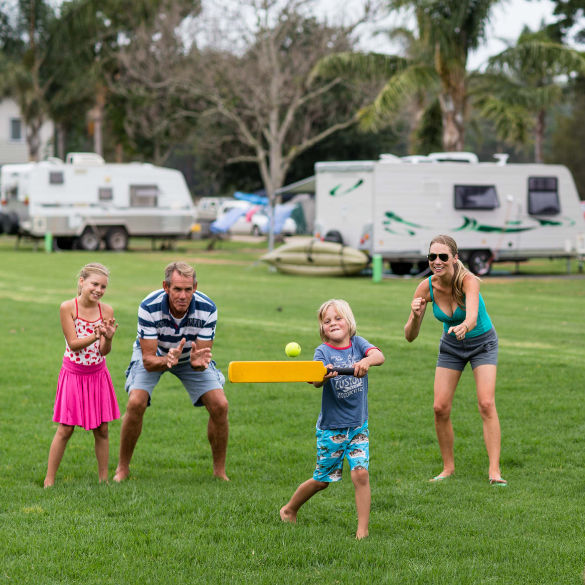 family playing cricket on grass with caravans in back ground