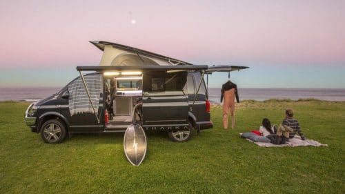 VW trakkadu parked on grass near ocean with surf board and wet suit