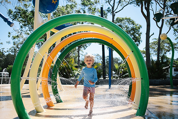 Smiling child running through rings spraying water at a water park at NRMA Holiday Parks Ocean Beach