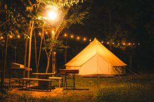 Night and warm light exposure of an illuminated bell tent surrounded by trees and a small camp fire at night, outdoor camping lifestyle