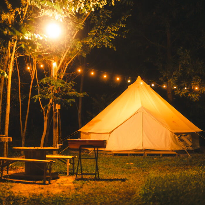 Night and warm light exposure of an illuminated bell tent surrounded by trees and a small camp fire at night, outdoor camping lifestyle
